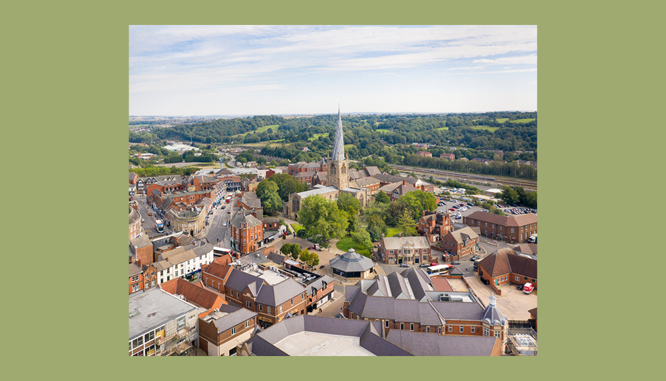 Aerial view of a town with a prominent church featuring a tall spire at its center, symbolizing community spirit and inclusive growth. Surrounding shops and houses blend harmoniously with the lush green countryside under a blue sky dotted with clouds.