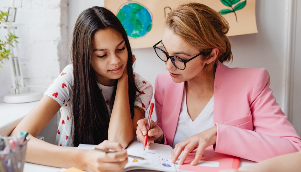 A woman in a pink blazer and glasses helps a girl with long dark hair on schoolwork at a table, fostering social connections. They discuss a notebook amid drawings, including a globe and a leaf.