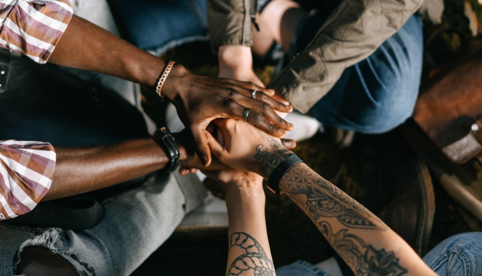 Four people sitting in a circle place their hands together in the center, symbolizing unity and teamwork. The image, rich with diverse skin tones and tattoos, highlights inclusivity, collaboration, and the strength built through social connections.