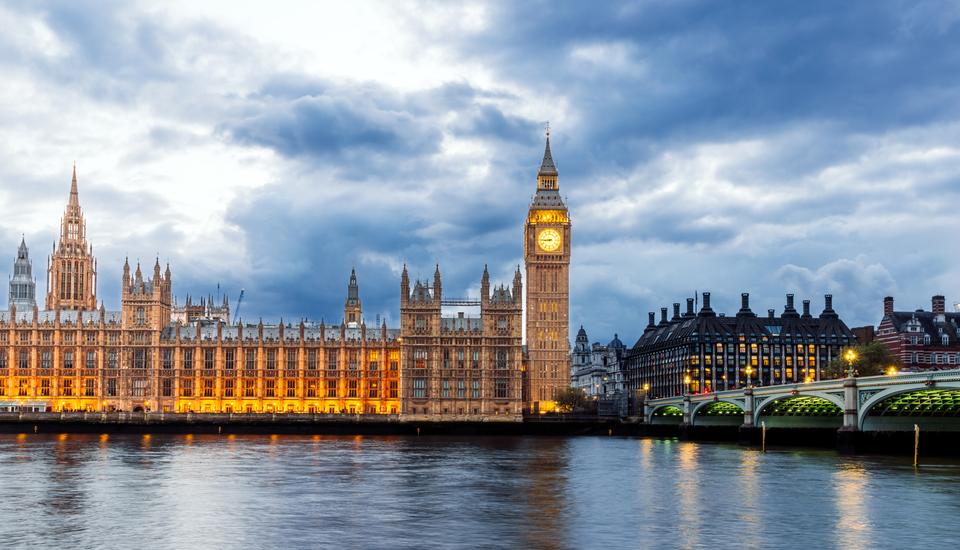 The image shows the Palace of Westminster and Big Ben illuminated at twilight. The buildings are reflected in the Thames River. The sky is cloudy, and Westminster Bridge is visible to the right.