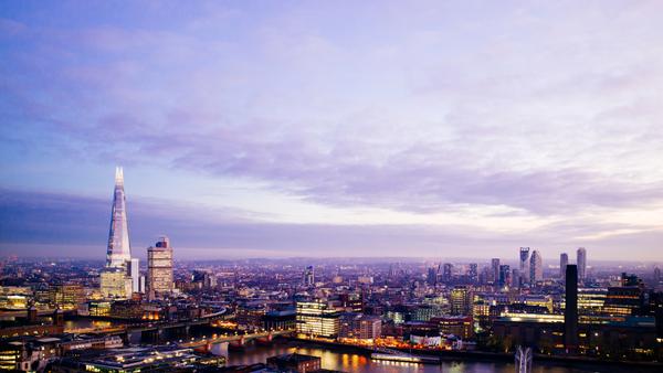 A panoramic view of a city skyline at dusk, featuring tall skyscrapers and the illuminated Shard building. The sky is a blend of purple and blue hues, with scattered clouds above the urban landscape.