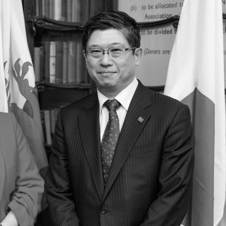 A man in a suit and tie stands indoors, smiling slightly. Behind him are flags and bookshelves. The image is in black and white. Join us to celebrate the opening of the UK Pavilion at Expo 2025 Osaka and hear from leading innovators.
