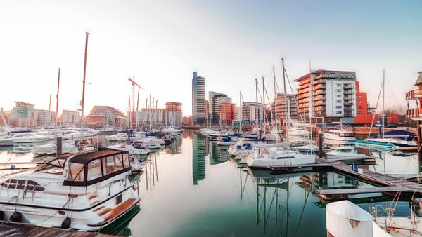 A serene marina scene at sunrise with numerous sailboats and yachts docked. Modern high-rise buildings and cranes line the waterfront. The calm water reflects the boats and skyline, creating a symmetrical view.