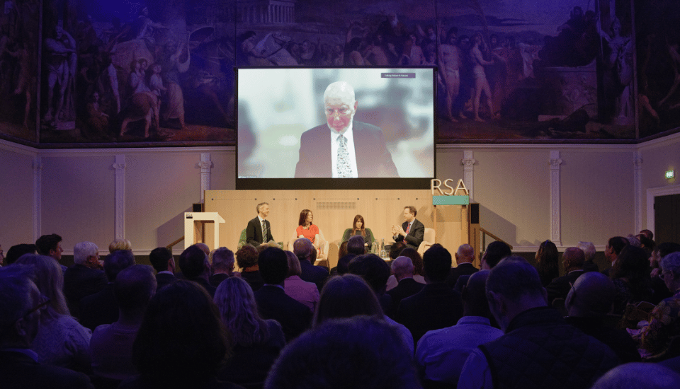 A diverse audience watches a panel discussion with three speakers on stage, while a large screen displays a fourth person appearing remotely. The room, adorned with classical wall art and the RSA logo, buzzes with discussions on social connection and insights inspired by Robert Putnam's work.