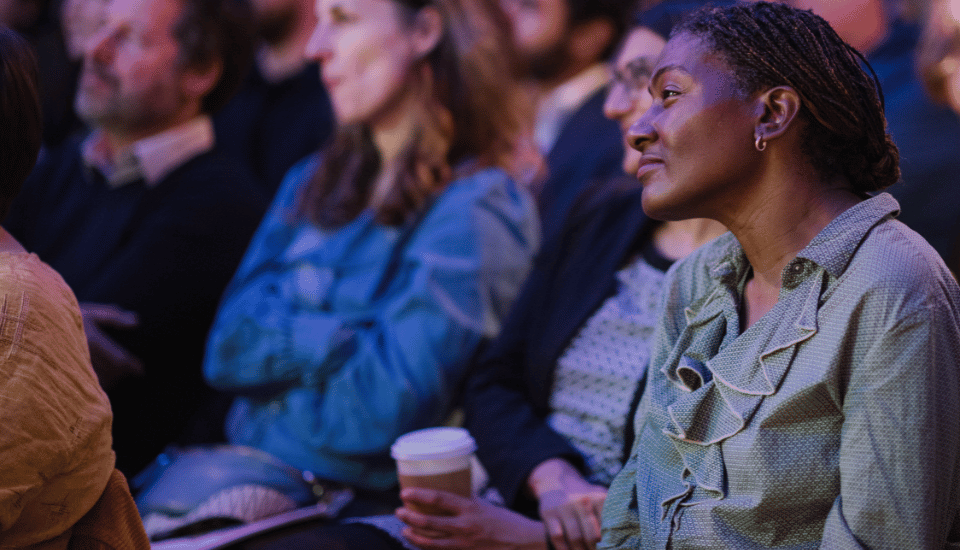 A person with braided hair, wearing a green blouse, is deeply engaged as they listen during an event on social capital. Holding a cup amid the audience, their attentive expression mirrors the dimly lit scene's focus on connection and community.