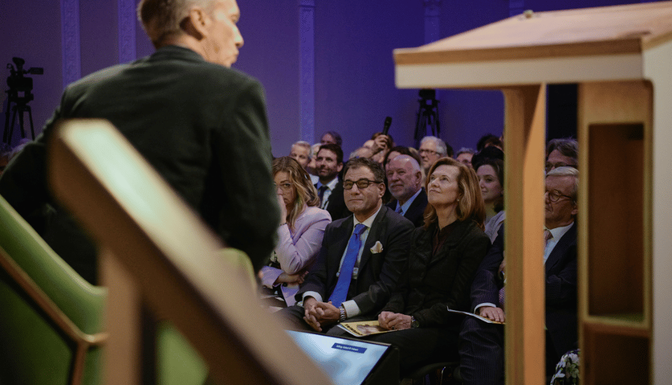 A speaker addresses an audience seated in rows. The listeners, including men and women, connect socially as they attentively watch, some taking notes. The room is illuminated with a blue tone, and a camera setup is visible in the background.