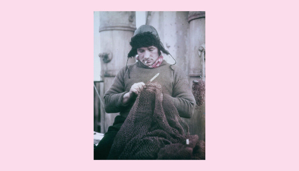 A person wearing a thick sweater, scarf, and ear-flapped hat is sitting and repairing a fishing net with a needle. Industrial equipment is visible in the background. The image has a vintage look with a pink border.