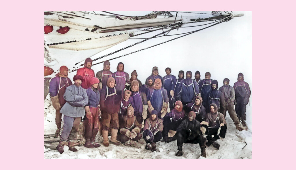 A group of people dressed in early 20th-century Antarctic exploration attire, standing optimistic on snow next to a ship's rigging and sails. They wear heavy coats, hats, and boots, set against a snowy landscape and pale pink border. Taken by photographer on  board Endurance