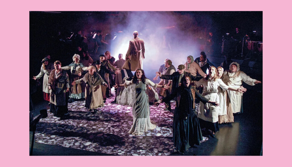 A group of performers on stage in a theatrical production, wearing period costumes. They stand under dramatic lighting with smoke effects, their arms outstretched. The atmosphere appears intense and dynamic.