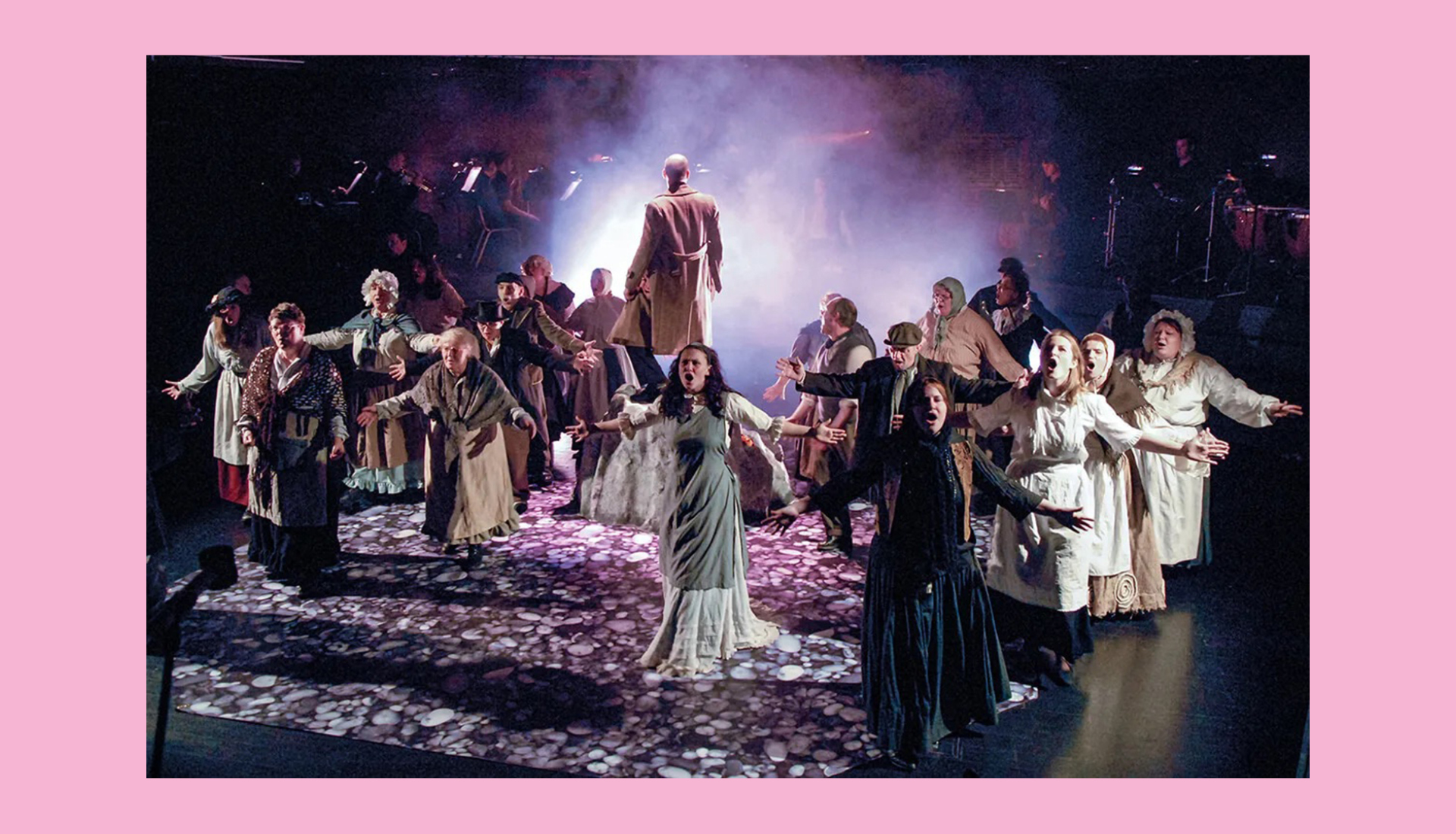 A group of performers on stage in a theatrical production, wearing period costumes. They stand under dramatic lighting with smoke effects, their arms outstretched. The atmosphere appears intense and dynamic.