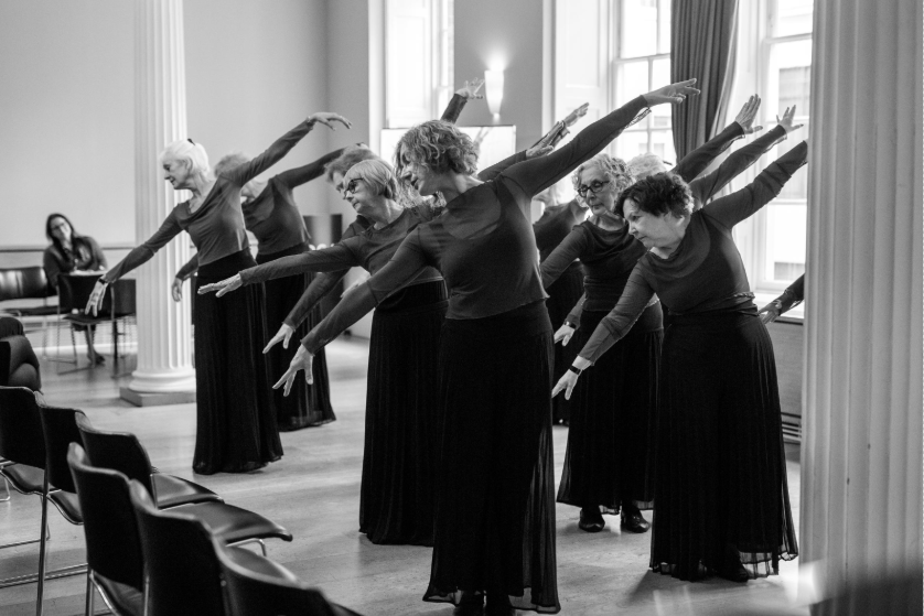A group of elderly women in long skirts and tops perform a synchronized dance in a spacious room with tall windows. They extend their arms gracefully in a forward-leaning position. A seated person observes from the back. Black and white image.
