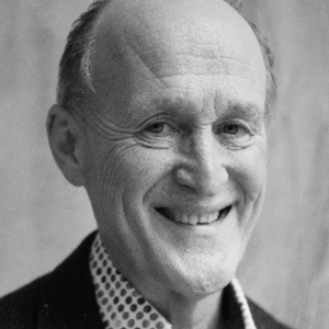 A grayscale portrait of an older man with a bald head and slight side hair, smiling, as he invites you to celebrate the opening of the UK Pavilion at Expo 2025 Osaka. He's wearing a dark blazer over a polka dot shirt. The background is softly blurred.