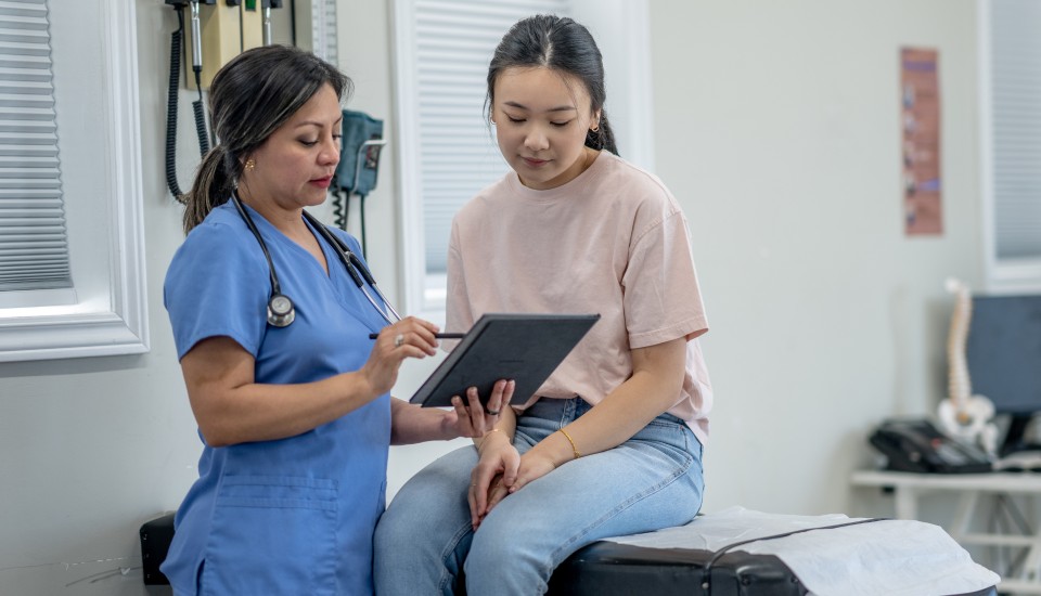 A healthcare professional in blue scrubs is demonstrating an AI-driven tablet to a patient sitting on the examination table. The patient, focused on women's health, wears a pink shirt and jeans. Medical equipment lines the walls in the background.