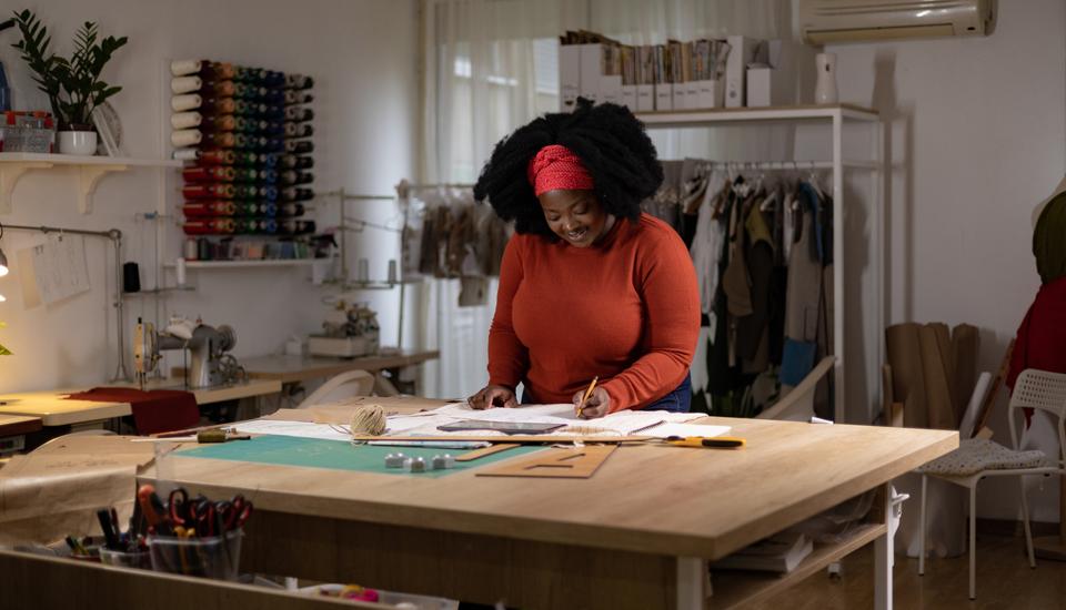 A smiling person works on fashion designs at a large table in a studio. They're using a pencil and paper while surrounded by sewing materials and clothing racks. The room is well-lit and organized.