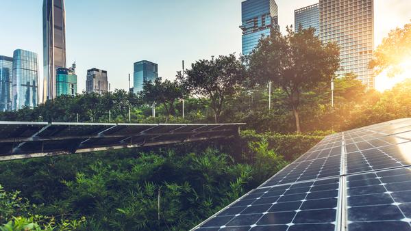 Solar panels in the foreground absorb sunlight, while green foliage surrounds them. In the background, modern skyscrapers rise against a clear sky, with the sun setting or rising, casting a warm glow over the urban scene.