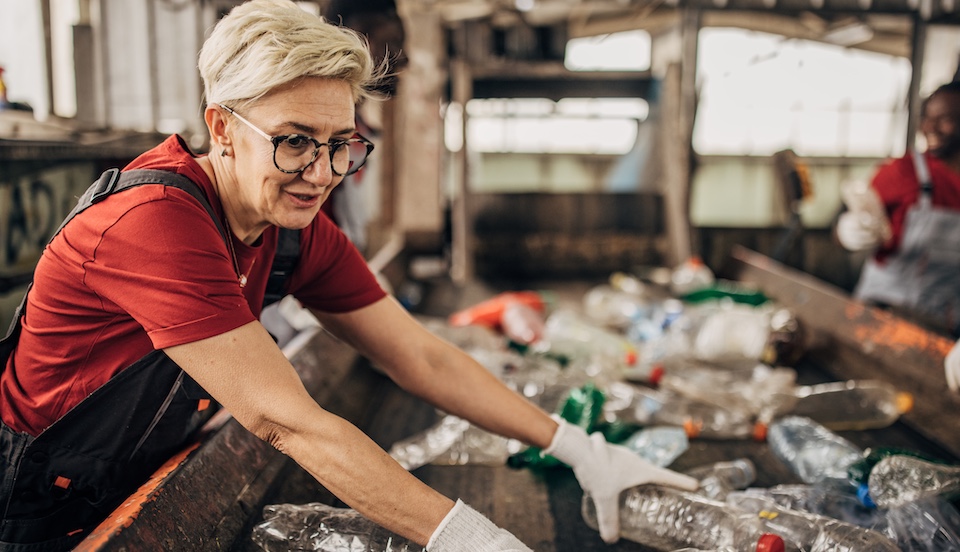 A person with short blond hair and glasses is sorting plastic bottles on a conveyor belt in a recycling facility. They are wearing a red shirt, dark overalls, and white gloves, working alongside other individuals in the background.