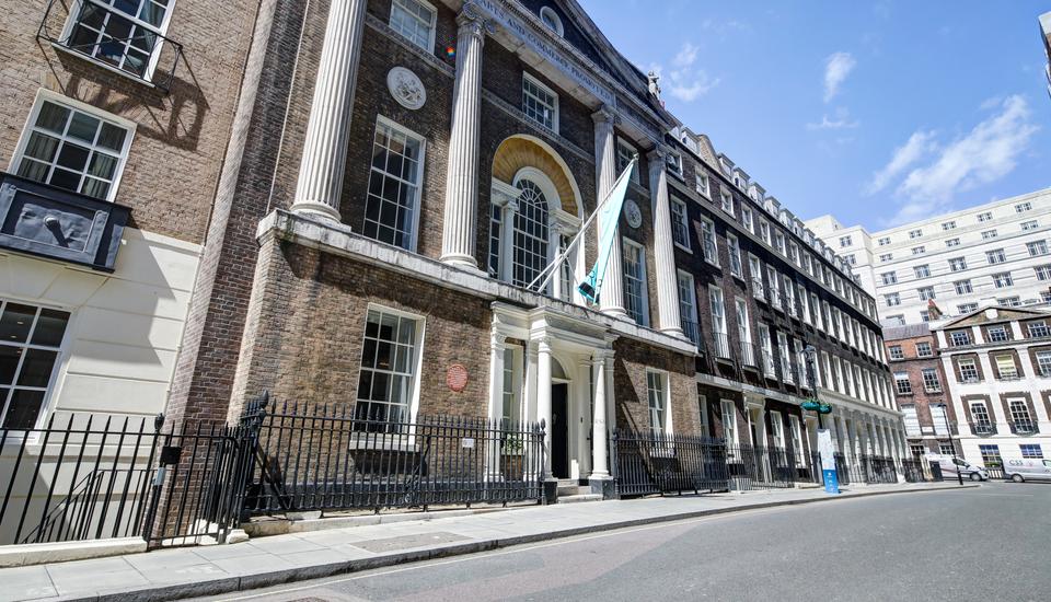 Street view of a historic building with a grand facade, featuring tall columns and large windows, reminiscent of where fellows might gather for a festival. It's encircled by a black wrought-iron fence beneath the clear blue sky, with neighboring structures visible on the right.