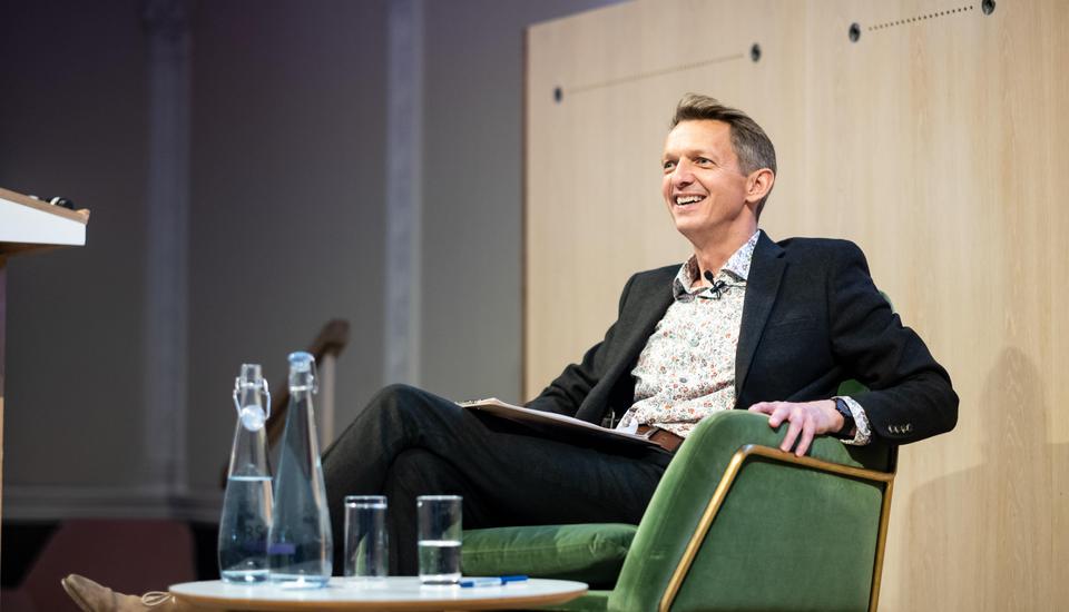 A smiling man in a suit sits in a green chair on stage, holding papers. A table with a water carafe and glasses is in front of him. He appears to be participating in a discussion or interview.