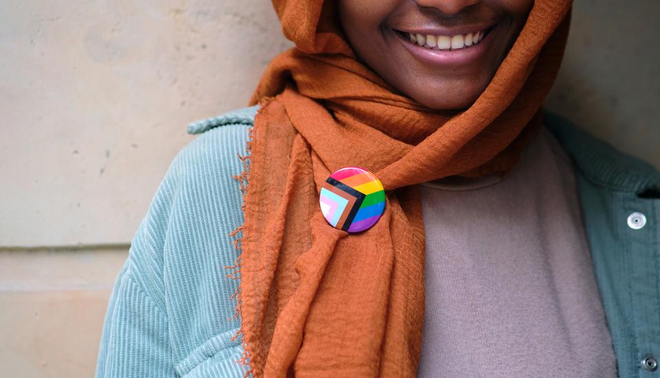 Smiling person wearing an orange hijab and green jacket, with a small pin displaying a rainbow and transgender pride flag design on the hijab.