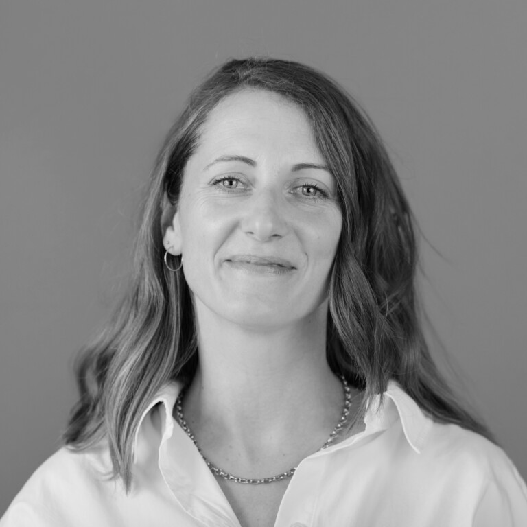 Black and white portrait of a woman with wavy hair, wearing a light-colored shirt, a necklace, and small hoop earrings, smiling gently with quiet trust in front of a plain background.
