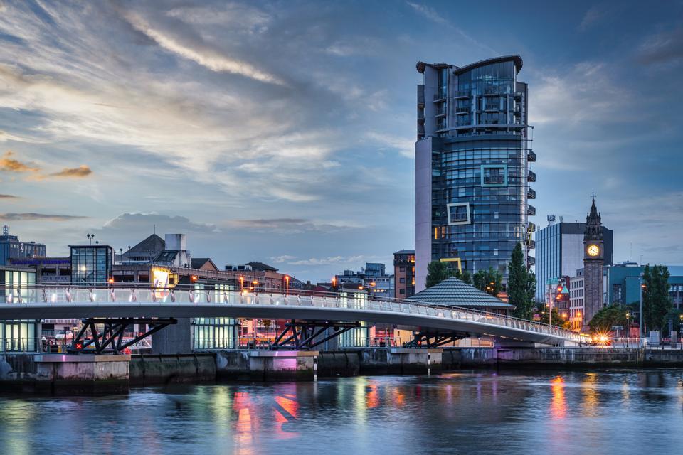 A modern pedestrian bridge spans over a calm river at dusk, leading to a cityscape with a tall, uniquely-shaped building. The sky is painted with soft clouds and hues of blue and orange, reflecting on the water below.