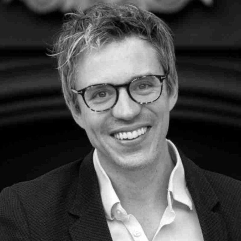 A man with light hair and glasses smiles at the camera, dressed in a light-colored shirt and dark jacket. The black and white image could be featured in promotions for sustainability events or UN Sustainable Development Goals initiatives.