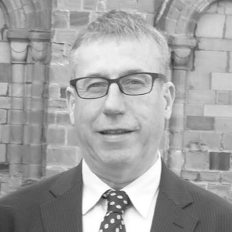 A middle-aged man wearing glasses and a suit with a polka dot tie stands in front of a stone building with arched windows. The image is in black and white.