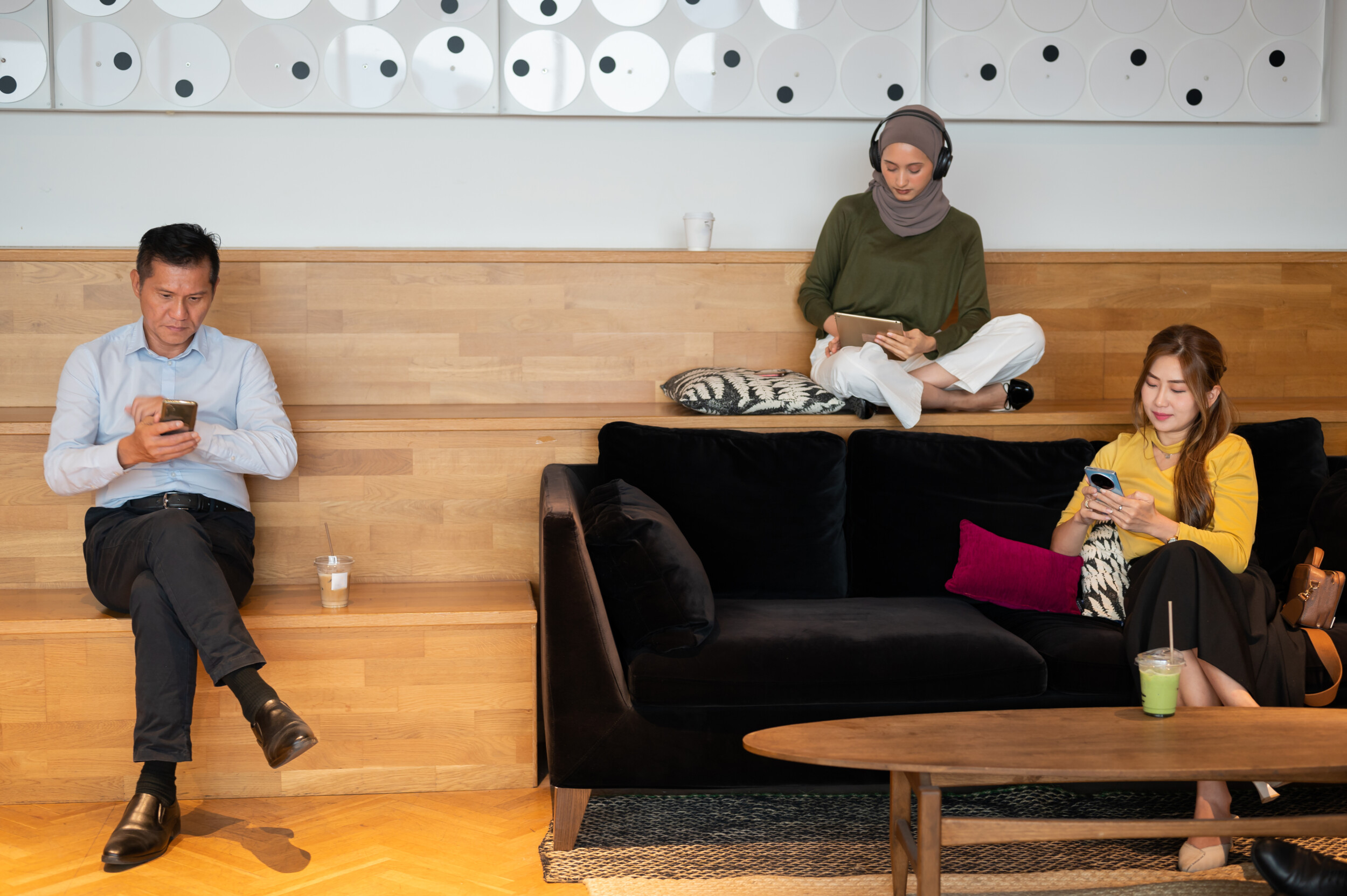 Three people are sitting in a modern lounge area. Two are on their phones, and one is using a tablet with headphones. They appear focused and relaxed, sitting on wooden steps and a black sofa with drinks nearby.