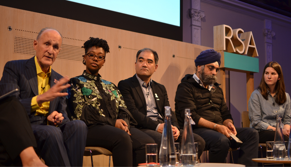 Five people sit on a panel discussion in front of an audience at the RSA, with microphones and water bottles on the table. The speakers appear attentive, discussing topics related to Expo 2025 as one person gestures while speaking.