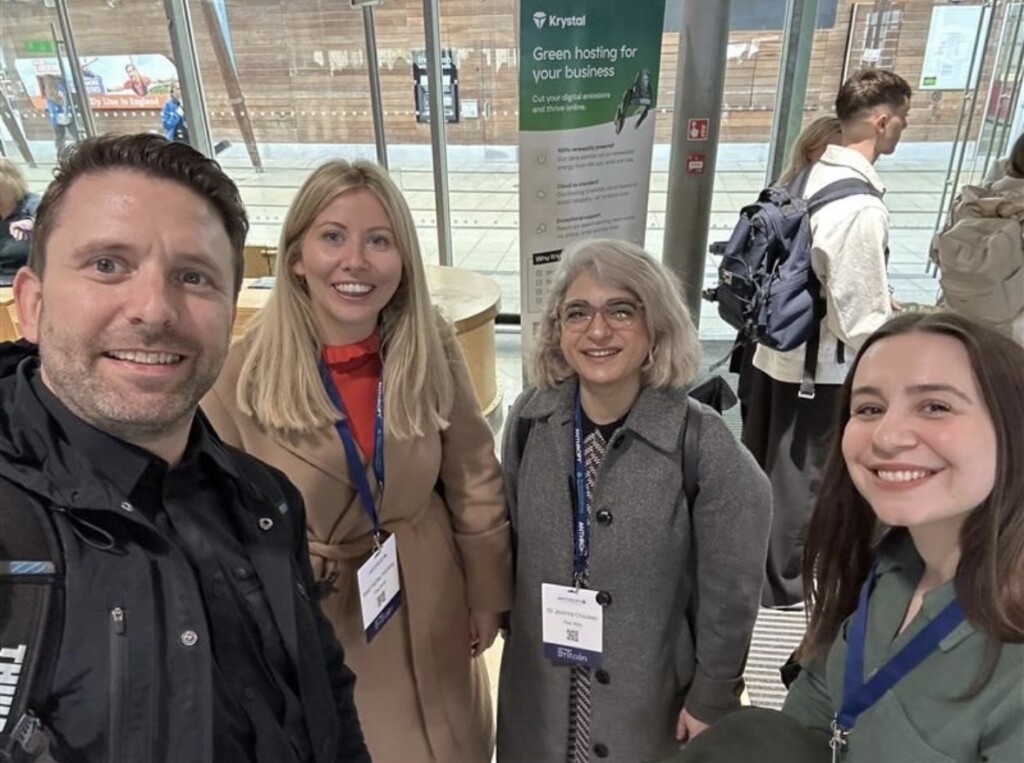Four people with conference badges, including RSA Trustees, smile for a selfie inside a modern building. Behind them, a poster reads “Green hosting for your business,” and several others are visible in the background.