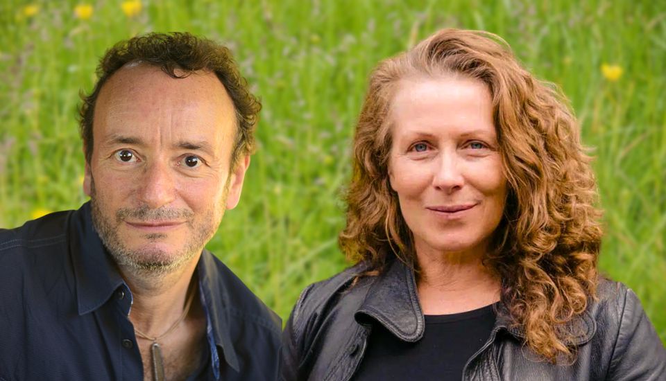 A man with short curly hair and a beard, wearing a dark shirt, and a woman with long curly hair, wearing a dark jacket, are smiling in front of a blurred grassy background.