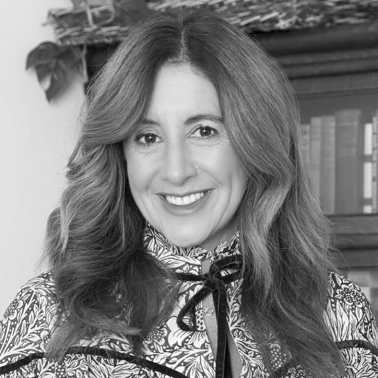 A woman with wavy, shoulder-length hair smiles at the camera. She is wearing a patterned top with a bow at the collar. There are books and a decorative basket in the background. The image is in black and white.