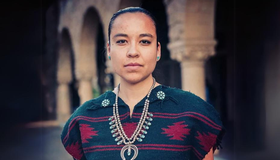 Person wearing traditional-style clothing, standing outdoors with arched stone architecture in the background. They have a serious expression, jewelry with turquoise stones, and their hair is pulled back.