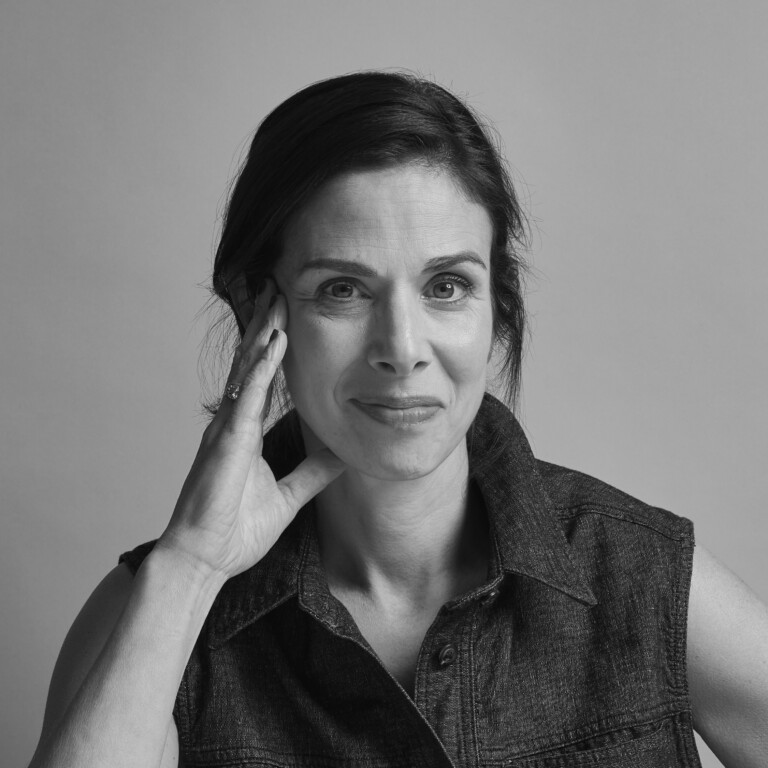 Black and white portrait of a woman with dark hair, wearing a sleeveless denim shirt, resting her head on her hand and smiling softly at the camera against a plain background.