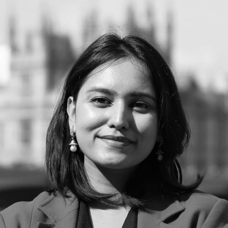 Black and white portrait of a woman with shoulder-length hair, wearing earrings and a blazer, smiling slightly. The blurred background hints at tall, pointed architectural structures reminiscent of the upcoming Osaka Expo 2025.