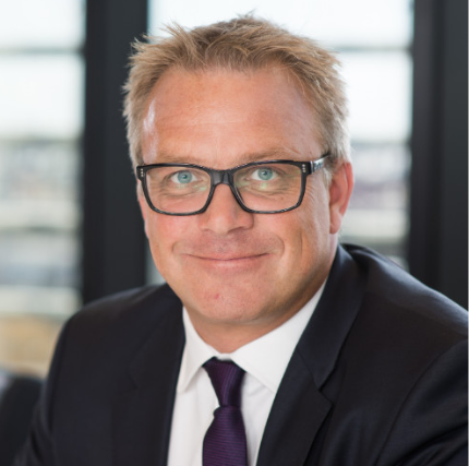 A man with light hair and glasses, dressed in a dark suit and tie, smiles warmly at the camera. He sits indoors, reminiscent of a fellows gathering, with a window softly blurred in the background.