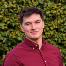 A young man with short dark hair, wearing a maroon button-up shirt, stands outdoors in front of a leafy green hedge, smiling slightly at the camera, embodying the approachable professionalism often seen among RSA trustees.