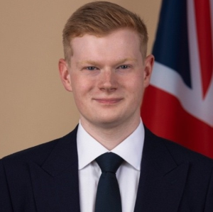 A young man with short red hair, dressed in a dark suit, white shirt, and dark tie, stands before a partial view of a Union Jack flag on a beige background, representing the professionalism of RSA Trustees.