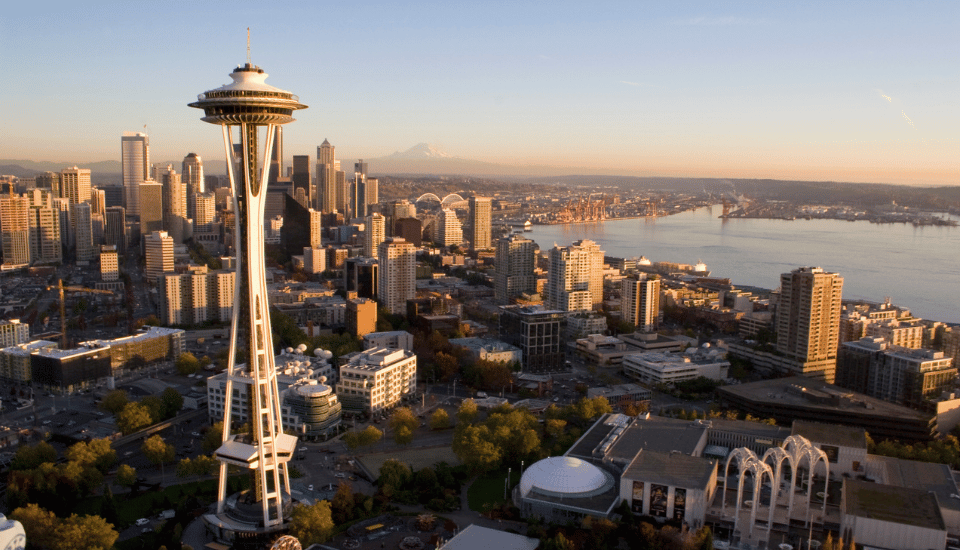 Aerial view of Seattle at sunset, where the iconic Space Needle stands as a symbol of social connection. The city skyline stretches toward the mountains and the waterfront, all bathed in a warm, golden hue under a clear sky.