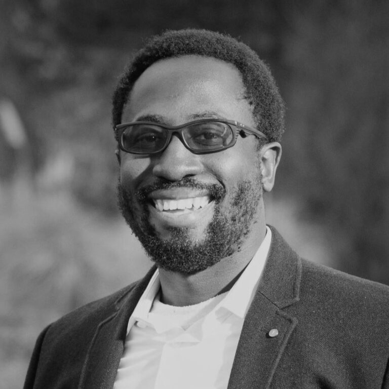 A man with short hair, a beard, and glasses smiles in a suit jacket over a white shirt. Captured in black and white with blurred outdoor scenery, he looks ready to lead the future of sustainability at a regeneration event.