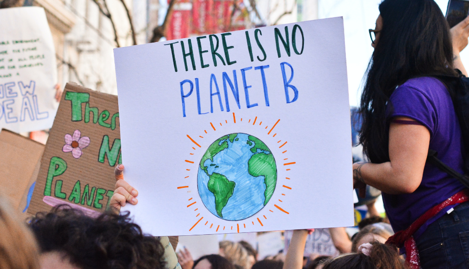 A person holds a sign at a climate protest that reads "There is no Planet B" above a drawing of the Earth. Other protest signs and people are visible in the background.
