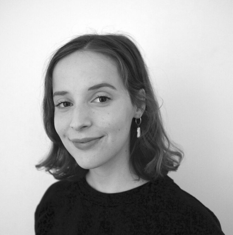 Tessa Devreese. A young woman with shoulder-length hair, wearing a dark top and hoop earrings, smiles softly while looking at the camera against a plain, light background. The photo is in black and white.