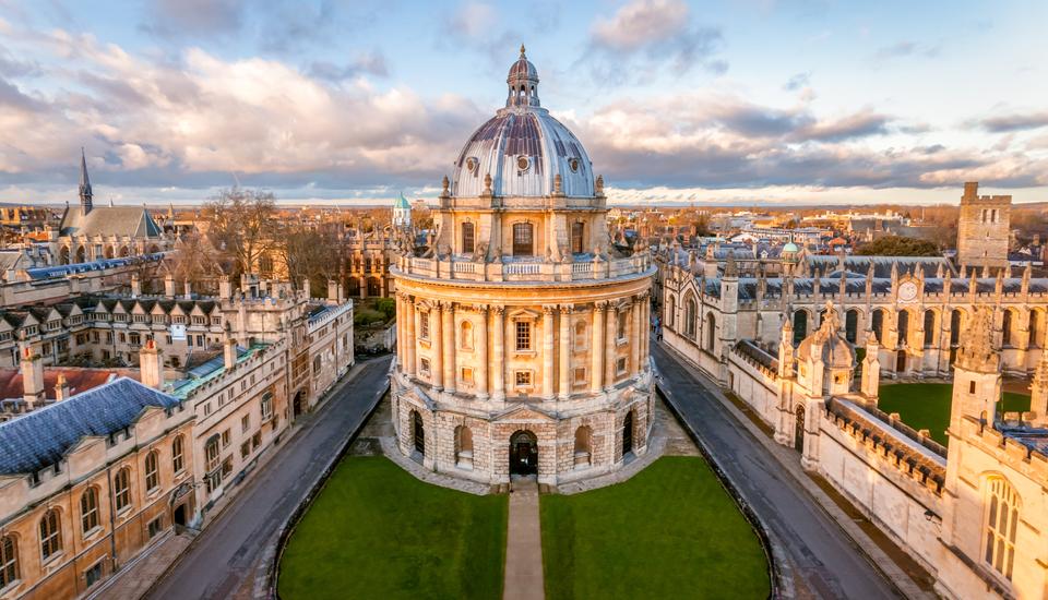 Aerial view of a historical cityscape in Oxford, England, featuring the iconic Radcliffe Camera. The round, domed building stands gracefully amid traditional sandstone structures and green lawns under a partly cloudy sky—a scene reminiscent of RSA's architectural appreciation.