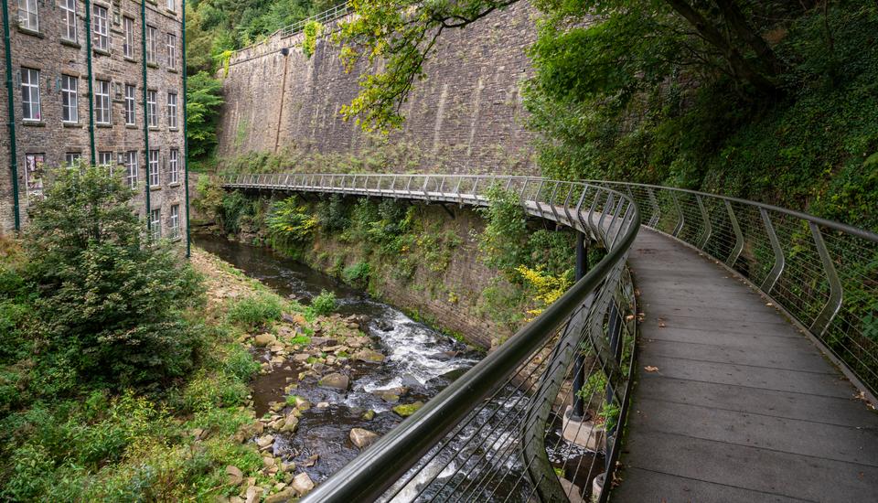 Curved metal walkway over a small river with rocky banks, bordered by lush greenery and trees. On one side, there's a tall brick building, and on the other, a stone wall runs parallel to the path.