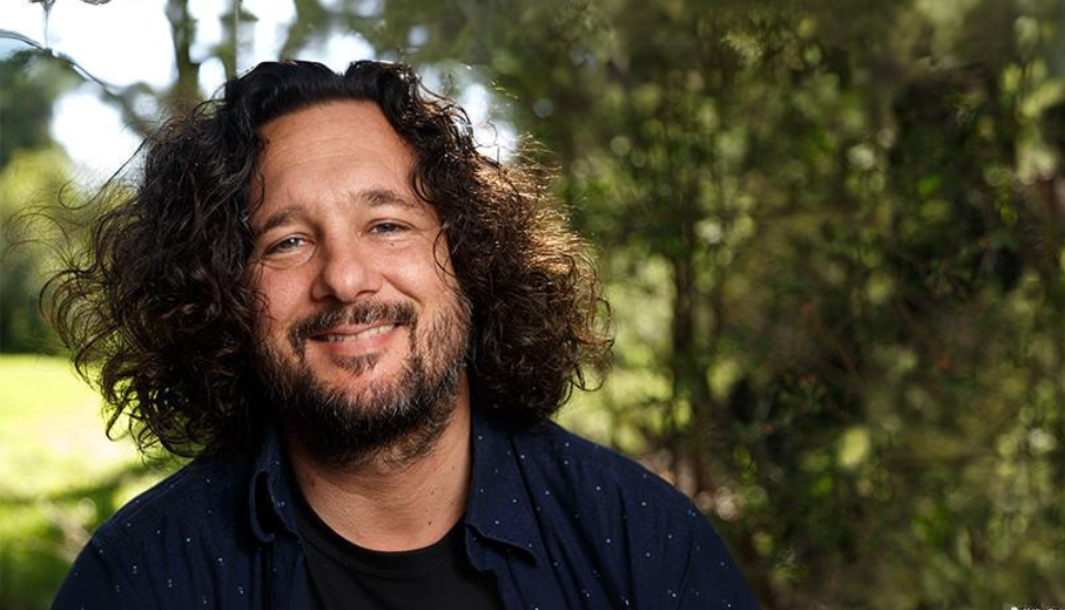 A person with curly hair and a beard is smiling while standing outside. They are wearing a dark shirt. The background features greenery with trees and foliage, suggesting a park or garden setting.