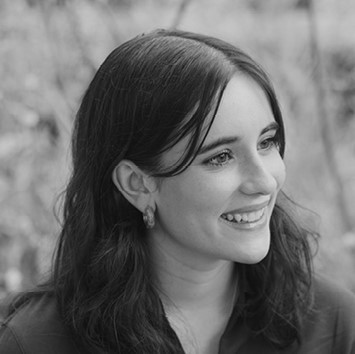 A young woman with dark hair and earrings smiles while looking to the side in an outdoor setting. The image is in black and white.