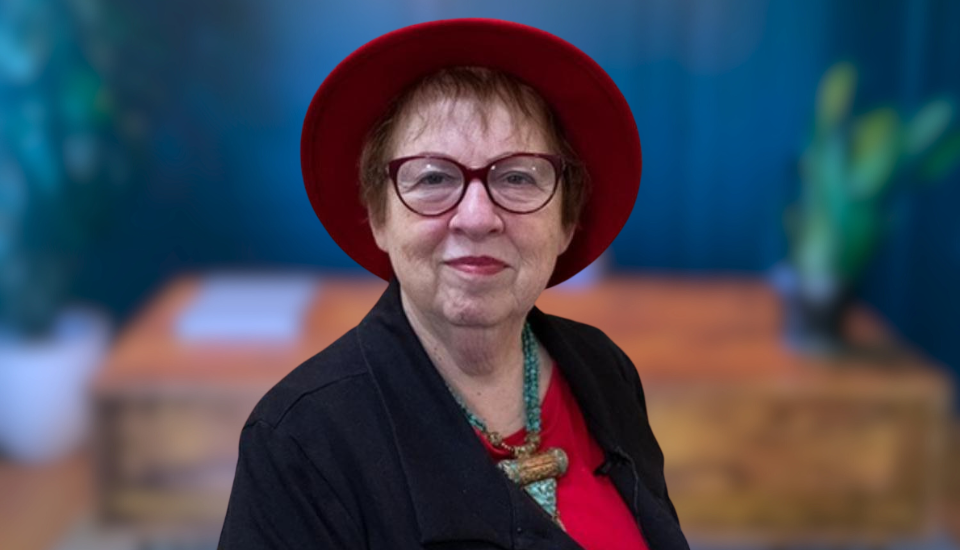 Elderly woman wearing a red hat, black jacket, and red shirt, smiling in front of a blurred office background with plants on each side.