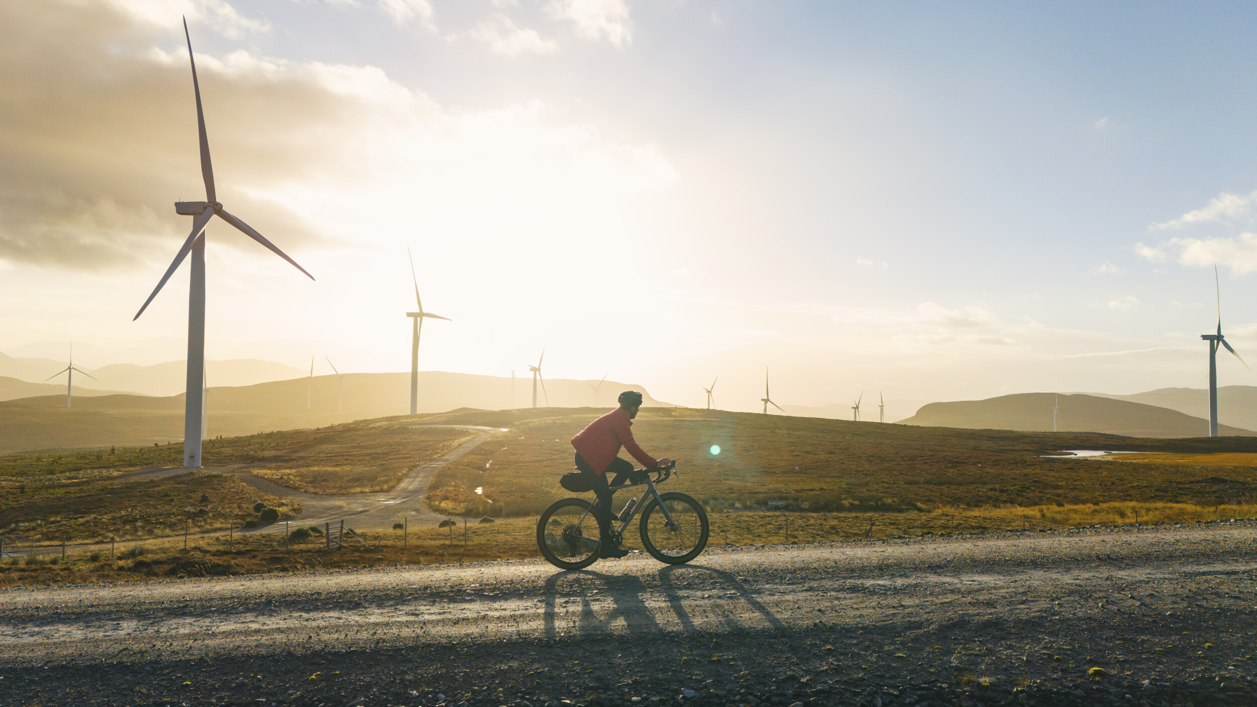 A person rides a bicycle on a gravel path in a vast landscape dotted with wind turbines. The sun is setting in the background, casting a warm glow over the scene. The sky is partly cloudy, creating a serene and sustainable energy theme.