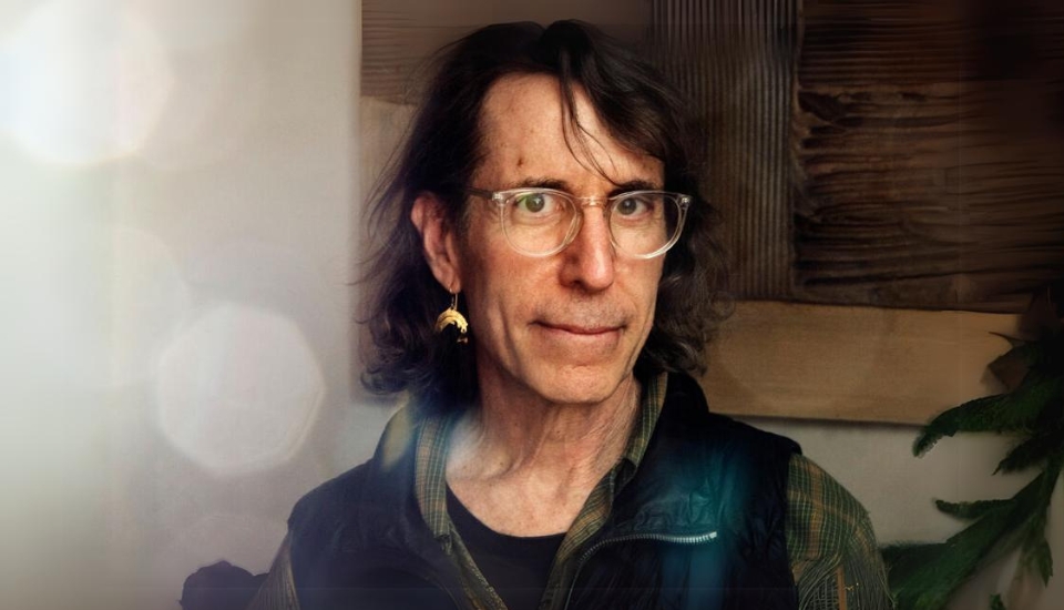 A person with long hair and glasses, wearing a plaid shirt and vest, looks at the camera. They have an earring and stand in front of a blurred background with a plant on the side.