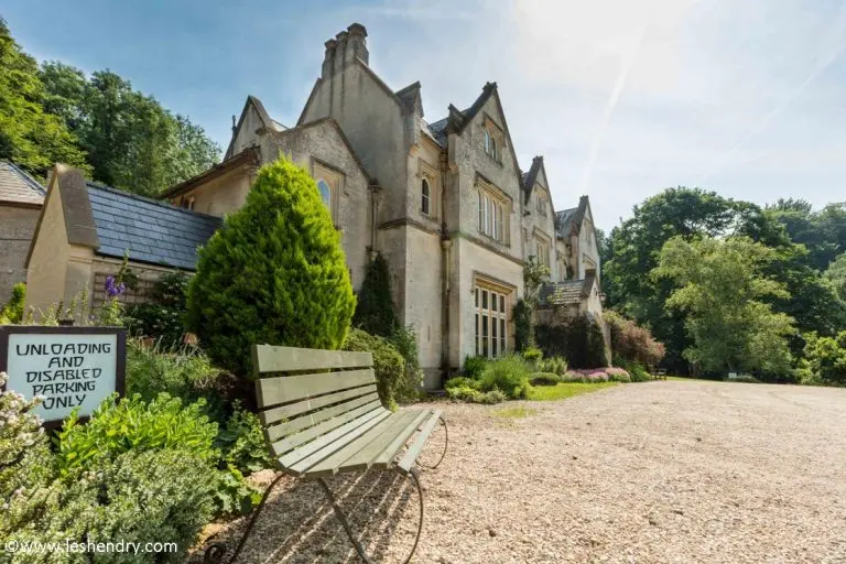 A large, historic stone house with steep gables sits among lush greenery and gardens. A green bench and a sign for unloading and disabled parking are visible on the gravel driveway in front.